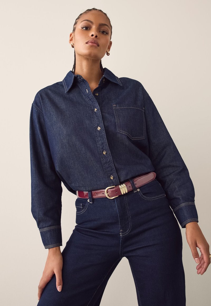 Jeanshemd in Dunkelblau mit durchgehendem Knopfleistenverschluss, langen Ärmeln und einer Brusttasche. Kombiniert mit hochtaillierter dunkler Jeanshose und einem burgunderroten Gürtel.