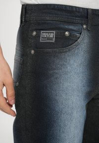 Temno modre kavbojke iz jeansa z bledenim gradientom, srebrno strojno opremo in etiketo Versace Jeans Couture na zadnjem žepu.
