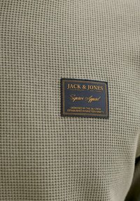 Ύφασμα μπεζ ζακάρ με μια μαύρη και χρυσή ορθογώνια ετικέτα ρούχων Jack & Jones ραμμένη στην περιοχή του στήθους.