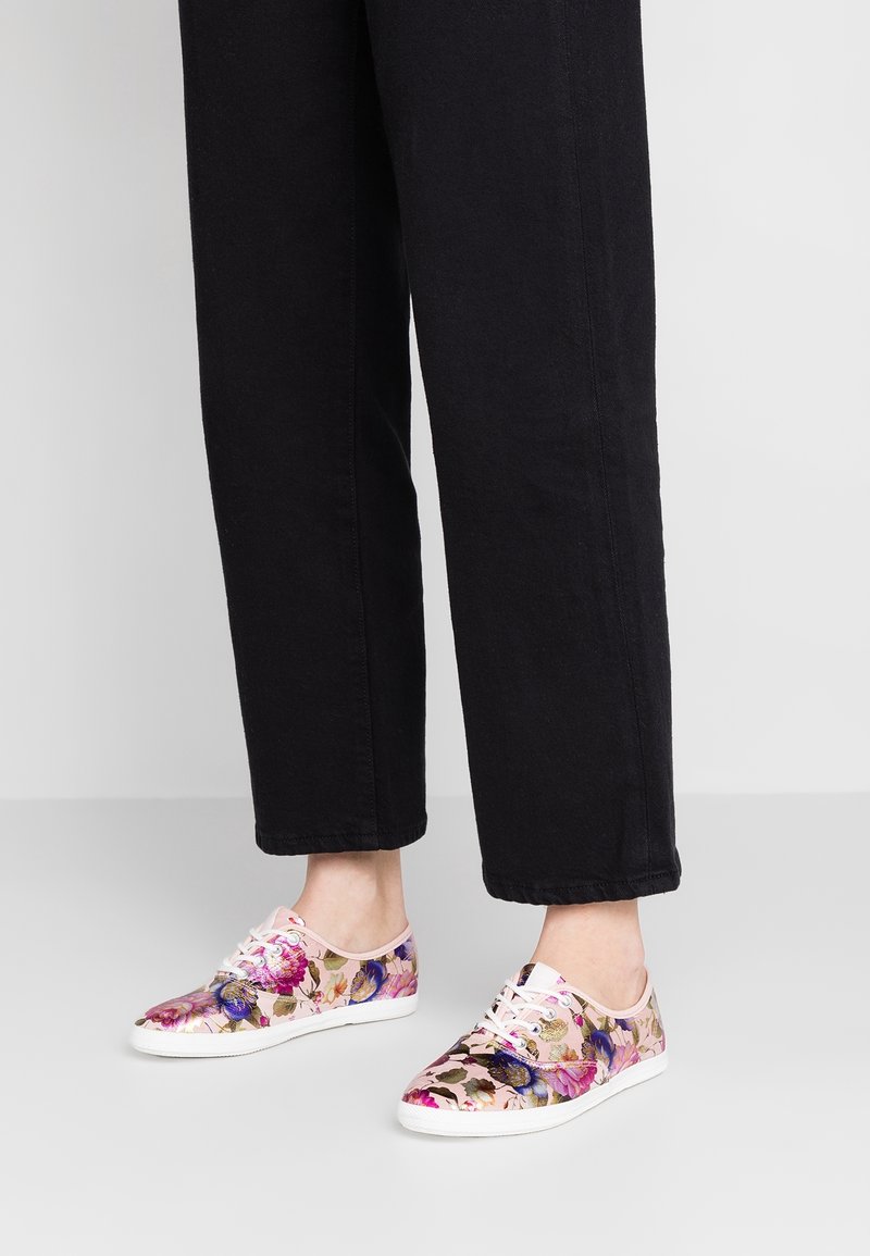 Jambes portant un pantalon noir et des baskets à lacets à motifs floraux colorés avec des semelles blanches sur un sol gris clair.