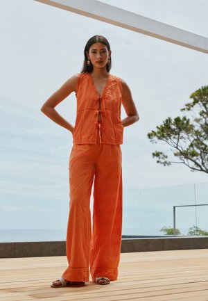 Naine seisab õues puidust terrassil, kandes varrukateta oranži toppi esiäärega ja sobivaid laiade säärtega pükse, pilvise taeva all.