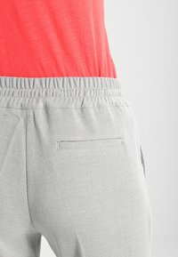 Pantalon gris texturé avec une ceinture élastique et une poche arrière, associé à un haut rouge. Le tissu présente un motif subtil.