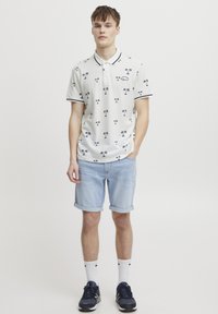 Hvit polo-skjorte med marineblå palme-motiv, svart stripete krage og mansjetter. Lyseblå rullet denimshorts, hvite sokker og mørke sneakers.