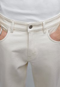 Pantaloni in denim beige con taglio dritto, dotati di tasche frontali, chiusura con bottone e anello metallico attaccato al passante della cintura.