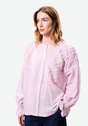 Femme aux cheveux ondulés mi-longs portant un chemisier rose pâle à boutons avec des détails en dentelle et des manches longues bouffantes, regardant légèrement sur le côté.