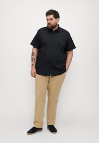 Zwarte korte mouwen overhemd, van katoen, gecombineerd met beige broeken en zwarte sneakers, met een relaxte pasvorm en zichtbare tatoeages op de arm.