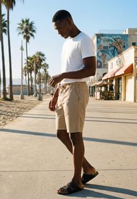 Bărbat în tricou alb, pantaloni scurți bej și sandale negre mergând pe o promenadă însorită de-a lungul plajei, marginită de palmieri, cu clădiri în fundal.