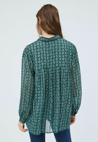 Genomskinlig grön blus med blommönster, rynkad detalj på baksidan, långa ärmar och rundad nederkant, vilket framhäver ett lätt tyg.