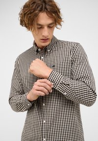 Junger Mann, der die Manschette seines schwarz-weiß-beigen karierten Hemdes an einem schlichten hellen Hintergrund anpasst.