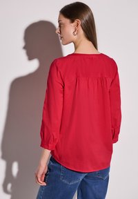 Rote Bluse mit langen Ärmeln, gerafftem Rückenteil und geknöpften Ärmeln, aus leichtem Stoff; kombiniert mit blauen Jeans.