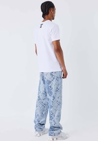 Maglietta bianca a maniche corte con testo nero sul retro, abbinata a jeans azzurri con motivo pixelato e scarpe da ginnastica bianche.