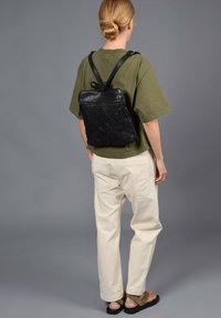 Sac à dos en cuir noir et en mesh avec des bretelles réglables, de forme rectangulaire et avec un haut zippé. Porté par un mannequin portant un pantalon clair et des sandales.