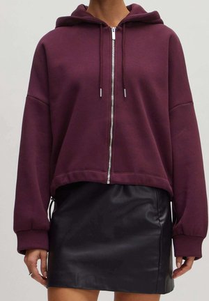 Vinrød hættetrøje med lynlås og snore, kombineret med en sort lædernederdel, båret af en stående person foran en ensfarvet baggrund.