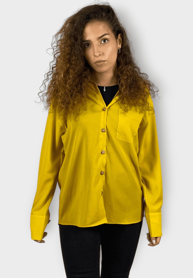 Camisa de manga larga y botones de tejido amarillo brillante. Cuenta con un único bolsillo en el pecho y botones de madera. Llevada sobre ropa negra.