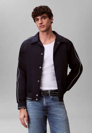 Jeune homme portant une veste noire avec un liseré blanc sur un débardeur blanc et un jean bleu, se tenant devant un fond gris uni.