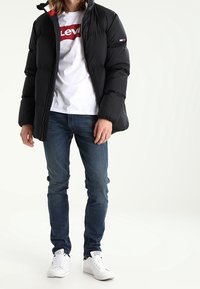 Schwarze Pufferjacke mit Reißverschluss, getragen über einem weißen T-Shirt mit rotem Logo. Kombiniert mit blauen Jeans und weißen Sneakern.