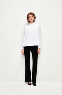 Blusa bianca a maniche lunghe con colletto arricciato e pieghe verticali, abbinata a pantaloni neri a vita alta e svasati, con tacchi a punta neri.