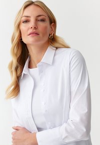Hvid skjorte med krave, lange ærmer og en glat tekstur, lagdelt over en hvid t-shirt. Har klare plastikknapper.