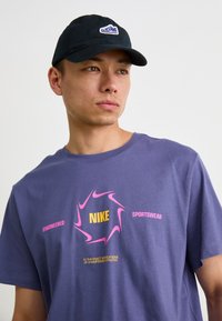Violet medvilnės marškinėliai su rožine ir geltona grafinėmis logotipais, kuriuose ant priekio yra užrašas "ENGINEERED SPORTSWEAR". Juoda kepurė su logotipu.