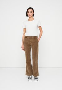 Καφέ παντελόνι κοτλέ με καμπάνα, υφή και εμπρόσθιες τσέπες, συνδυασμένο με εφαρμοστό λευκό μπλουζάκι και μαύρα αθλητικά παπούτσια.