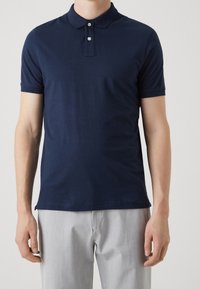 Mann in einem taillierten, marineblauen Polohemd mit kurzen Ärmeln und zwei weißen Knöpfen, kombiniert mit hellgrauen Hosen, vor einem schlichten Hintergrund stehend.