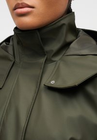 Κοντινό πλάνο ενός ατόμου που φοράει μια αδιάβροχη χακί καμπαρντίνα με γιακά ψηλού τύπου και λεπτομέρεια με πατιλέτα και κουμπιά στον ώμο.