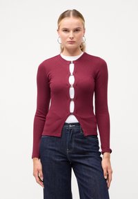 Cardigan lung cu maneci lungi, de culoare bordeaux, cu nervuri, dotat cu accesorii decorative din argint și un decolteu tăiat, purtat peste un top alb și blugi de denim închisi la culoare.