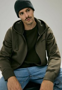 Brązowa bluza z zamkiem błyskawicznym i teksturowanym kapturem, w połączeniu z ciemnozieloną koszulą i niebieskimi dżinsami. Ciemna czapka beanie dopełnia outfit.