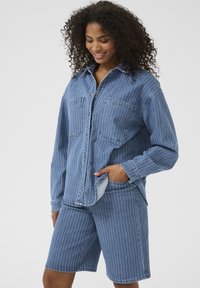 Denimist särk, millel on vertikaalsed valged triibud, kaks rinna taskut, nööbiga disain, koos sobivate denim lühikesed pükste, mis on samuti triibulised.