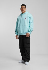 Lyseblå hettegenser med frontlomme og rødt hjerte-logo, kombinert med svarte cargo-bukser og hvite joggesko. Stoffet ser mykt og avslappet ut.