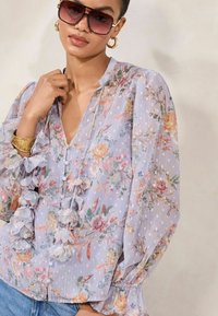 Bluză florală transparentă în lavandă deschisă, cu mâneci fluturate, nasturi și decolteu în V; prezintă diverse modele colorate de flori și buline.