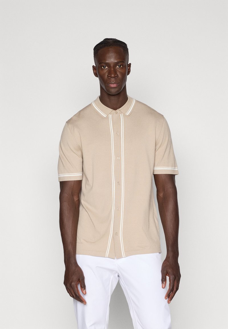 Šviesiai rudos spalvos trumpomis rankovėmis polo marškiniai su sagomis ir kontrastingomis baltomis juostelėmis apykakle ir rankogaliais. Lygus paviršius.