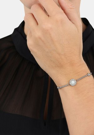Bracciale in argento con un charm rotondo al centro in perla, catena texturizzata e piccoli accenti decorativi. Indossato su una mano contro un tessuto scuro.