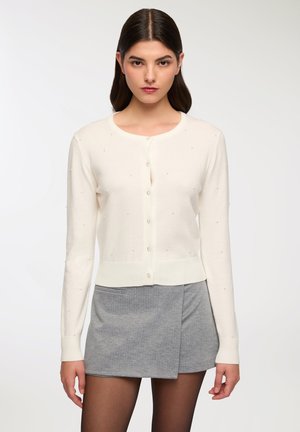 Cardigan bianco lavorato a maglia con maniche lunghe, scollo rotondo e chiusura a bottoni. Presenta piccole decorazioni testurizzate su tutta la superficie.