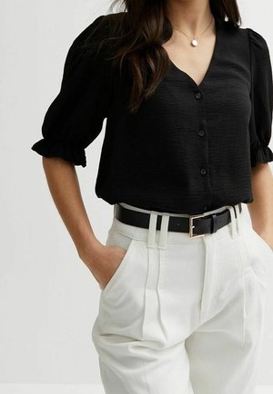 Kvinde iført sort bluse med knapper og pufærmer, hvide højttaljede bukser med plissering, sort bælte og vedhængshalskæde, hænderne i lommerne.