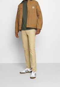 Brown canvas jacket with two front pockets, green t-shirt, beige trousers, and white sneakers with black stripes.