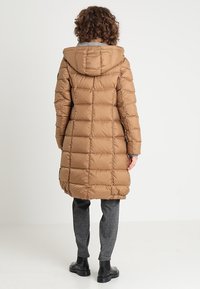 Abrigo acolchado marrón con capucha, que presenta un diseño acolchado, de largo y una textura suave y brillante. Lleva puesto sobre pantalones grises con estampado y botas negras.