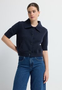 Navyblaue, kurz geschnittene Strickjacke aus weichem Material mit großem Kragen, kurzen Ärmeln und Knopfverschluss. Verfügt über Vordertaschen und gerippten Saum.