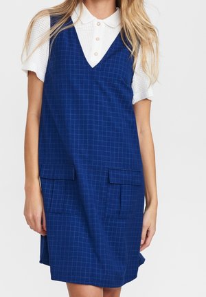 Kvinde iført en blå ternet ærmeløs kjole med frontlommer over en hvid kortærmet poloshirt, vist fra skuldre til knæ.