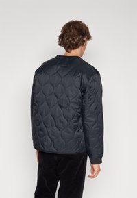 Veste matelassée rembourrée en noir uni avec un motif texturé en forme de vagues. Elle dispose d'un col rond et de poignets côtelés. Matériau doux.