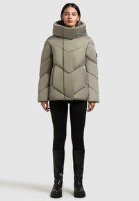 Puffjacke in hellolivgrün mit hohem Kragen, Zickzack-Steppung und Reißverschlusstaschen. Kombiniert mit schwarzen Leggings und kniehohen Stiefeln.