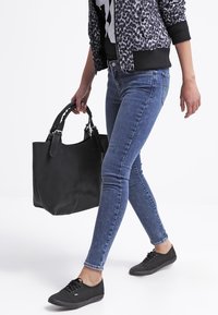 Bolso de cuero negro con asas cortas. Llevado con jeans ajustados azules y zapatillas negras. Modelo con una chaqueta bomber estampada.