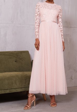 Femme portant une longue robe en dentelle florale rose clair avec des manches transparentes et une jupe plissée, debout à côté d'un canapé vert.