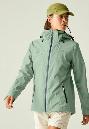 Femme portant une veste imperméable vert clair et une casquette vert olive, regardant sur le côté, avec un short beige sur un fond uni.