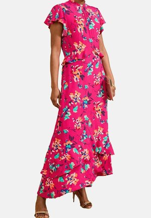 Femme portant une robe maxi fleurie rose vif avec des manches volantées, un ourlet à volants, et tenant une petite pochette.