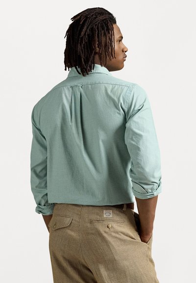 Cămașă mint green cu mâneci lungi și cu mânecile suflecate, având un pliu la spate, asociată cu pantaloni maro cu carouri. Accent pe textura materialului și culoare.