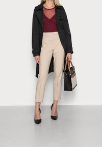 Svart trenchcoat över en vinröd topp med volangkant, tillsammans med beige cigarrettbyxor och svarta spetsiga klackar, hållande en svart handväska.