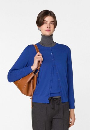 Frau trägt eine blaue, geknöpfte Strickjacke über einem grauen Rollkragenpullover, dunkle Hose, trägt eine braune Leder-Umhängetasche.