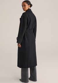 Cappotto trench in lana nera con cintura, maniche lunghe e dettagli dei polsini con bottoni. Abbinato a pantaloni grigi a gamba larga con un design minimalista.