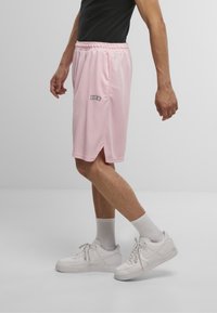 Lyserøde træningsshorts med sideslids, elastisk talje og sort logo-detalje på venstre ben. Båret med hvide sneakers og strømper.
